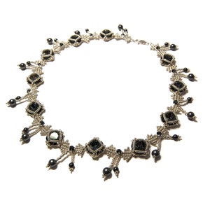 May include: A silver and black beaded necklace with a geometric design. The necklace features black beads and silver beads arranged in a pattern of squares and triangles. The necklace has a silver clasp.