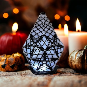 May include: A white and black geometric lantern with a light inside. The lantern is sitting on a wooden surface in front of a blurred background of candles and pumpkins.