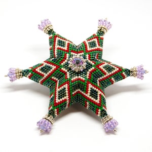Puede incluir: Un adorno de estrella de cuentas verdes, rojas y blancas con un cristal morado en el centro. La estrella tiene un patrón geométrico y está decorada con pequeñas cuentas moradas.