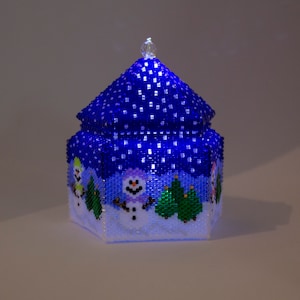 May include: A blue beaded Christmas ornament shaped like a house with a white snowy base and three snowmen. The house has a white star on top and is lit from within.