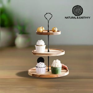 May include: A three-tiered wooden serving stand with a black metal central support. The stand holds cupcakes and chocolate candies. The top tier has two candies, the middle tier has a cupcake, and the bottom tier has two cupcakes. The stand is branded with the text "NATURAL & EARTHYY".