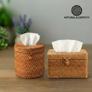 May include: Two woven rattan tissue box holders, one round and one rectangular, each with white tissues protruding from the top. The tissue boxes are a warm brown color and have a natural, rustic aesthetic. The brand name "NATURAL & EARTHYY" is visible.