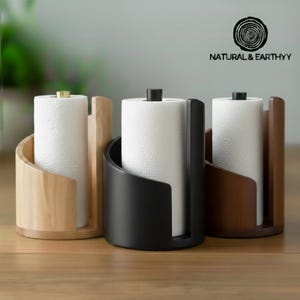 May include: Three paper towel holders in natural wood, black, and brown. Each holder has a curved design and holds a white paper towel roll. The holders are on a wooden surface. The logo "NATURAL & EARTHYY" is in the upper right corner.