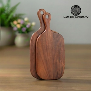 Op de afbeelding: Twee houten snijplanken met handvatten. De planken zijn diepbruin van kleur en hebben een gladde afwerking. De handvatten hebben een gat om op te hangen. De planken zijn van Natural & Earthyy.