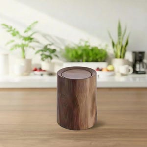 Può includere: Un cestino cilindrico in legno con un coperchio piatto e circolare. Il cestino è realizzato in legno marrone scuro con un motivo a venature naturali. Il cestino è su una superficie di legno marrone chiaro, con piante e oggetti da cucina sullo sfondo.