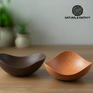 May include: Two wooden bowls with a wave-like design. One bowl is a dark brown color, and the other is a light brown color. The bowls are sitting on a wooden surface. The text "NATURAL & EARTHYY" is in the upper right corner.