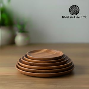 Puede incluir: Una pila de seis platos de madera de diferentes tamaños, dispuestos de mayor a menor. Los platos son de color marrón cálido y parecen ser de madera natural. El texto "NATURAL & EARTHYY" es visible en el fondo.