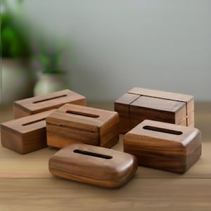 May include: Several wooden tissue box holders in various shapes and sizes. The boxes are made of a warm-toned wood and feature a rectangular opening on top for tissue dispensing. The boxes are arranged on a light brown wooden surface.