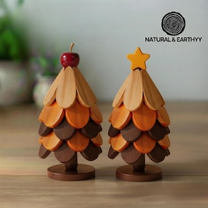 Pode incluir: Duas esculturas decorativas de árvores em madeira com camadas de escamas de madeira em forma de pétalas em tons de castanho claro, médio e escuro. Uma árvore é rematada com uma pequena maçã vermelha, a outra com uma estrela amarela. O texto NATURAL & EARTHYY é visível.