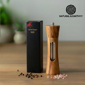 May include: A wooden salt and pepper mill with a clear viewing window, next to a black box labeled "Salt & Pepper Mill". The mill is made of light brown wood and sits on a wooden surface with peppercorns and pink salt.
