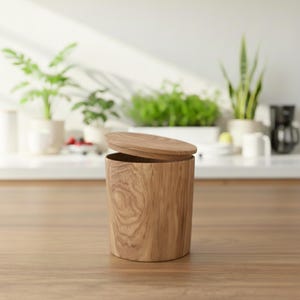 Op de afbeelding: Een lichtbruine houten container met een deksel, met de natuurlijke houtnerf. De cilindrische container staat op een houten oppervlak, met een heldere achtergrond met planten en keukenartikelen. Het deksel staat iets open.