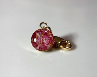 May include: A circular, gold-coloured charm featuring a pink and white floral pattern set in clear resin. It's connected to a gold-coloured clasp. This charm is a decorative accessory.