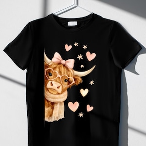 Può includere: T-shirt nero con un'illustrazione di una mucca delle Highlands. La mucca indossa occhiali, un fiocco e una sciarpa, con cuori e stelle rosa. La mucca è marrone e beige, con un fiocco e occhiali rosa.