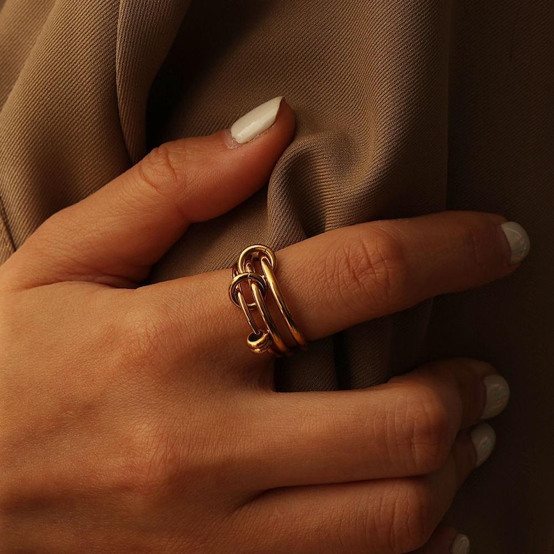 May include: A gold ring with a distinctive design of interlocking loops. The ring is displayed on a finger, emphasizing its detailed craftsmanship and polished surface. The jewellery item is set against a plain backdrop, accentuating its elegant shape.