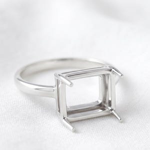 May include: A polished silver-toned metal ring setting featuring a rectangular head with four prongs designed to hold a gemstone. The band is smooth and circular, connecting to a raised, open-frame basket structure that is ready for a stone setting.