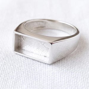 Könnte beinhalten: Ein silberfarbener Ring mit einer glatten, polierten Ringschiene, die in eine markante, rechteckige Fassung übergeht. Die Mitte der rechteckigen Fassung ist vertieft und weist eine texturierte, matte Oberfläche auf, die einen Kontrast zum glänzenden Metall bildet.