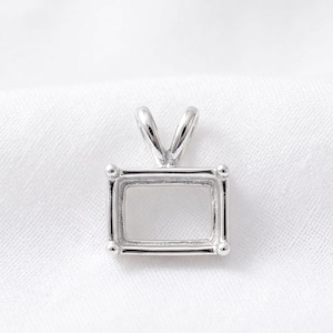 May include: A silver-toned rectangular pendant setting featuring a hollow center and a top bail for attachment. The metal frame has a smooth, polished finish with four small prongs at each corner to secure a stone or insert within the rectangular opening.