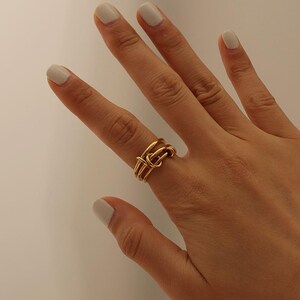May include: A gold ring with a knot design. The ring has multiple bands that interweave to form the knot. The ring is worn on a finger with light-coloured nail polish.