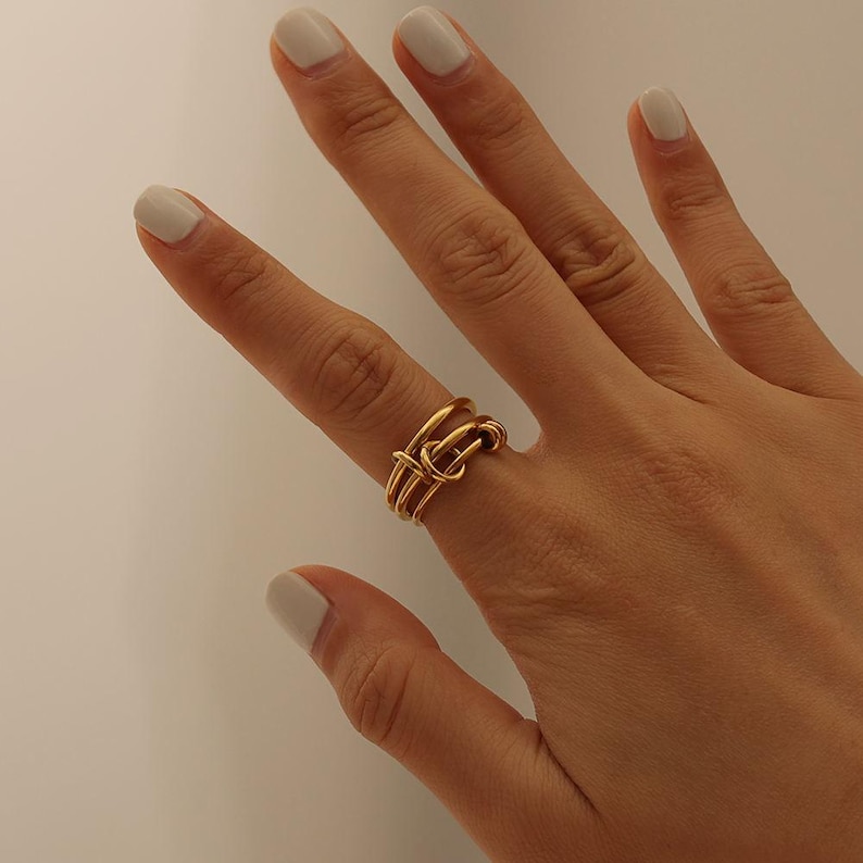 May include: A gold ring with a knot design. The ring has multiple bands that interweave to form the knot. The ring is worn on a finger with light-coloured nail polish.