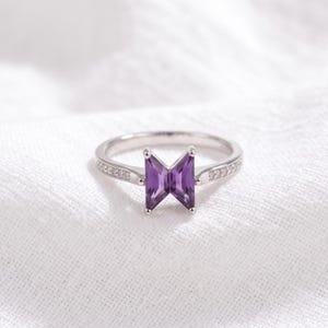 May include: A silver-toned ring featuring a central purple gemstone cut into a geometric, shield-like shape. The band is slender and accented with small, clear stones embedded along the sides, creating a refined and polished appearance.