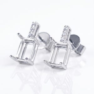 May include: A pair of silver-toned stud earrings. Each earring features a rectangular setting with four prongs and a vertical bar adorned with small, clear stones. The earrings are designed to hold gemstones.