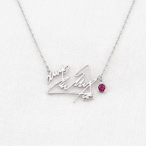 Puede incluir: Un collar de tono plateado con una cadena fina y un colgante central diseñado como una firma en cursiva. Una pequeña piedra preciosa redonda de color rosa vibrante está engastada y cuelga del lado derecho del colgante de metal.