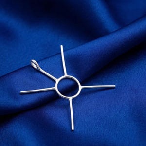 Pode incluir: Um pingente prateado com um centro circular e quatro linhas retas que se estendem para fora, mais um laço. O pingente está sobre um fundo de tecido azul escuro texturizado. O design é simples e geométrico.