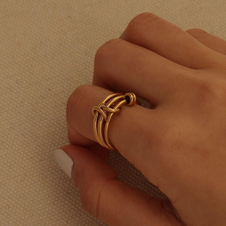 May include: A gold ring with an unusual design. The ring has multiple bands that cross and loop around each other, forming an intricate pattern. The ring is worn on a finger.