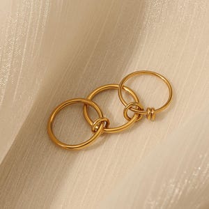 May include: A gold ring featuring three interlinked circles. The rings are joined with a knot design. The jewellery is presented on a cream-coloured, textured surface.