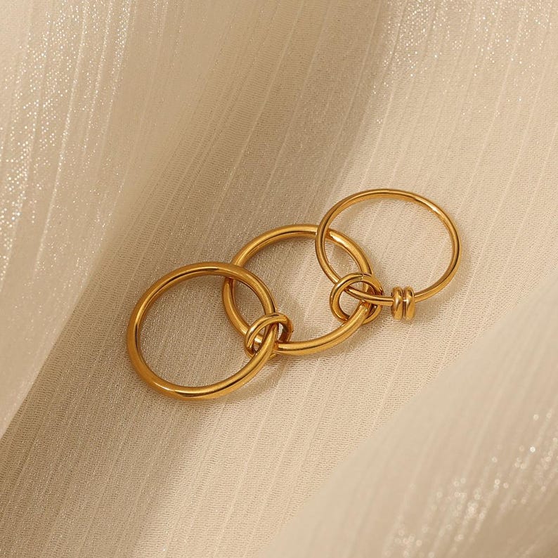 May include: A gold ring featuring three interlinked circles. The rings are joined with a knot design. The jewellery is presented on a cream-coloured, textured surface.