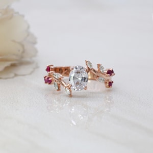 Peut inclure: Bague en or rose ornée d'une pierre précieuse ovale et transparente en son centre. La monture présente un motif de feuilles et de branches, agrémenté de petites pierres précieuses rouges et transparentes. La bague est posée sur un fond blanc doux.