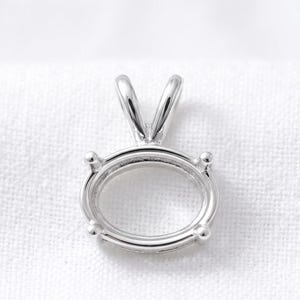 May include: A silver-toned metal pendant setting featuring an oval-shaped frame with four prongs designed to hold a gemstone. The setting includes a V-shaped bail at the top for attachment to a chain, showcasing a polished, reflective metallic finish.