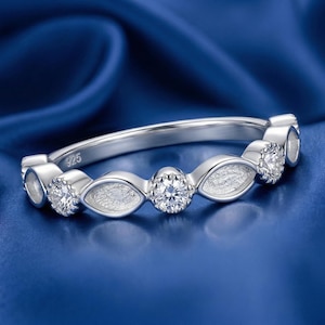 May include: A silver ring with a series of clear, round gemstones and marquise-shaped accents. The ring is set against a deep blue background. The band is marked with "925".