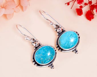May include: A pair of silver-toned dangle earrings featuring oval, turquoise-colored stones. The stones are set in a decorative frame with small, round accents. The earrings have a hook closure and are displayed on a light pink surface.