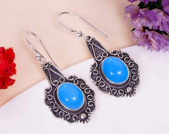May include: A pair of silver-toned drop earrings with a light blue oval stone center. The earrings have an ornate, swirling design around the stone and a hook closure. The background includes purple and red flowers.