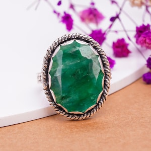 Könnte beinhalten: Ein silberner Ring mit einem großen, facettierten, smaragdgrünen Edelstein. Der ovale Edelstein ist in einer silbernen Lünette mit einem gedrehten Seilmuster gefasst. Der Ring wird auf einer weißen Oberfläche mit lila Blumen im Hintergrund präsentiert.