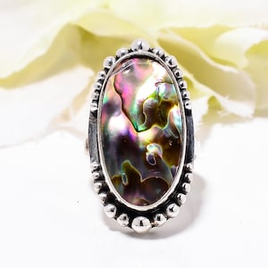Puede incluir: Anillo ovalado con una piedra central de concha de abulón iridiscente. La concha muestra una mezcla de colores, incluyendo verde, morado y dorado. El anillo tiene un engaste plateado con un borde de cuentas.