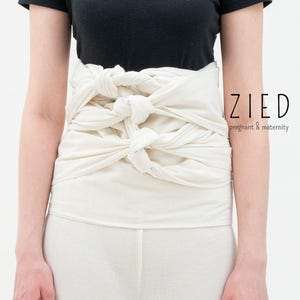 Puede incluir: Faja abdominal de maternidad de color crema con múltiples secciones anudadas. La faja se usa alrededor del abdomen, con el texto "ZIED pregnant & maternity" visible en el lado derecho.