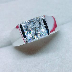 Puede incluir: Un anillo de tono plateado con una gran piedra transparente de corte cuadrado montada en estilo tensión. La banda es lisa y pulida con un acabado metálico reflectante. El texto RadhaJewelllers es visible en la esquina superior izquierda.