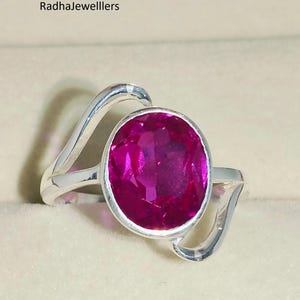 Könnte beinhalten: Ein silberner Ring mit einem großen, oval geschliffenen, leuchtend rosa Edelstein. Der Ring hat ein einzigartiges, fließendes Design, das sich um den Edelstein windet. Der Ring ist vor einem neutralen Hintergrund platziert, um die Farbe des Edelsteins und das Design des Rings hervorzuheben.