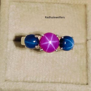 Op de afbeelding: Een zilveren ring met drie edelstenen. De middelste steen is magenta met een stervormig patroon, geflankeerd door twee diepblauwe, ronde edelstenen. De ring is geplaatst tegen een neutrale achtergrond.