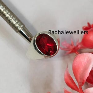 Puede incluir: Un anillo de plata con una gran piedra preciosa roja oscura de talla ovalada. El anillo está engastado en una banda de plata pulida y sencilla. También hay una herramienta plateada y acentos florales rojos, con el texto "RadhaJewelllers" visible.