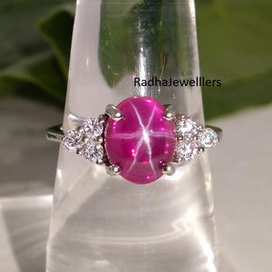 May include: A silver ring featuring a large, oval, star ruby gemstone. The ruby is a vibrant pink with a six-ray star pattern. Small clusters of clear gemstones flank the ruby on either side. The text "Radhajewelllers" is visible.