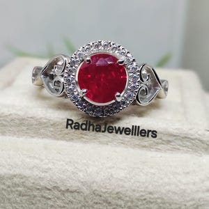Può includere: Un anello d'argento con una grande gemma rossa brillante e rotonda, circondata da pietre più piccole e chiare. L'anello ha una fascia decorativa con dettagli a filigrana a forma di cuore. Il testo "RadhaJewellers" è visibile.