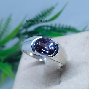Op de afbeelding: Een zilveren ring met een grote, ovale, donkerpaarse edelsteen. De ring heeft een eenvoudig, modern ontwerp met een gepolijste afwerking. De tekst "Radhajewelllers" is zichtbaar op de achtergrond.