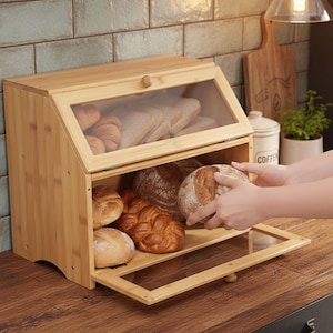 May include: A wooden bread bin with a clear glass door, filled with various types of bread. The bin is open, revealing loaves of bread and pastries. The bread bin is on a wooden surface.