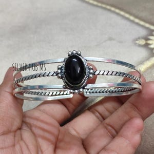 May include: A silver cuff bracelet with a large, oval black stone at the center. The bracelet has a three-band design, with a twisted rope pattern on the middle band. The text "SILVER HUB MS" is visible on the left side of the bracelet.