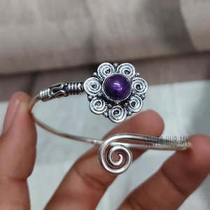 Può includere: Bracciale a polsino argentato con un elemento centrale a forma di fiore con una gemma viola scuro. Il bracciale presenta un design a spirale e dettagli intricati attorno alla gemma.