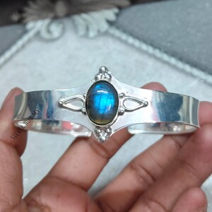 Puede incluir: Un brazalete de plata con una piedra preciosa ovalada azul iridiscente. El brazalete tiene elementos decorativos alrededor de la piedra preciosa y está estampado con "SILVER HUB MS". El brazalete es una banda ancha, diseñada para ser usada en la muñeca.
