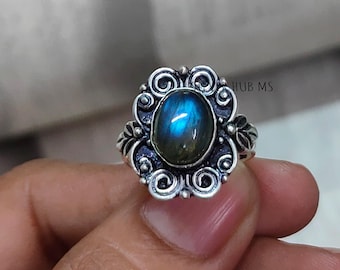 May include: A silver ring featuring an oval, iridescent blue gemstone. The gemstone is set in a decorative, filigree-style setting with swirling and floral details. The ring is held by a hand.
